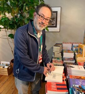 Photo of Benjamin Lefebvre, a forty-something white man with dark hair, dark glasses, and a grey beard, wearing a blue coat and beige pants, smiling for the camera and holding a purple pen in his left hand as he signs a copy of his book, /In the Key of Dale/, surrounded by piles of books and flanked by a leafy plant behind him.