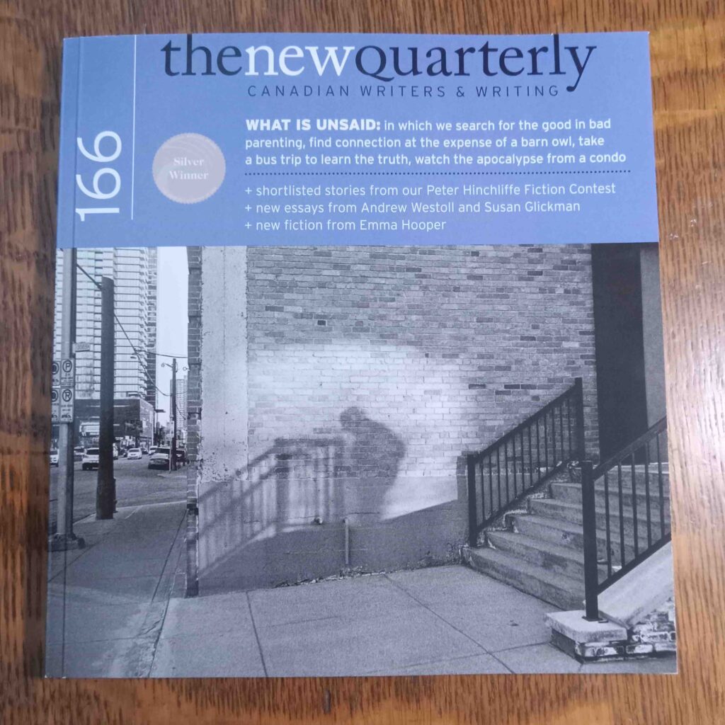 Copy of issue 166 of The New Quarterly against a brown wooden surface. The cover consists of a black and white photo of the shadow of a person checking their phone in an undisclosed outdoor location. The photo appears below the following text against a blue background: "The New Quarterly / Canadian Writers & Writing / 166 / WHAT IS UNSAID: in which we search for the good in bad parenting, find connection at the expense of a barn owl, take a bus trip to learn the truth, watch the apocalypse from a condo / + shortlisted stories from our Peter Hinchliffe Fiction Contest / + new essays from Andrew Westoll and Susan Glickman / + new fiction from Emma Hooper."